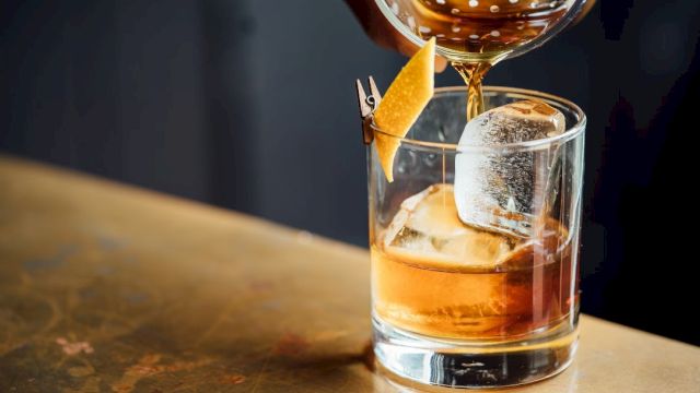 A short glass with whiskey and large ice, garnished with an orange twist, being poured a second time from a bottle.