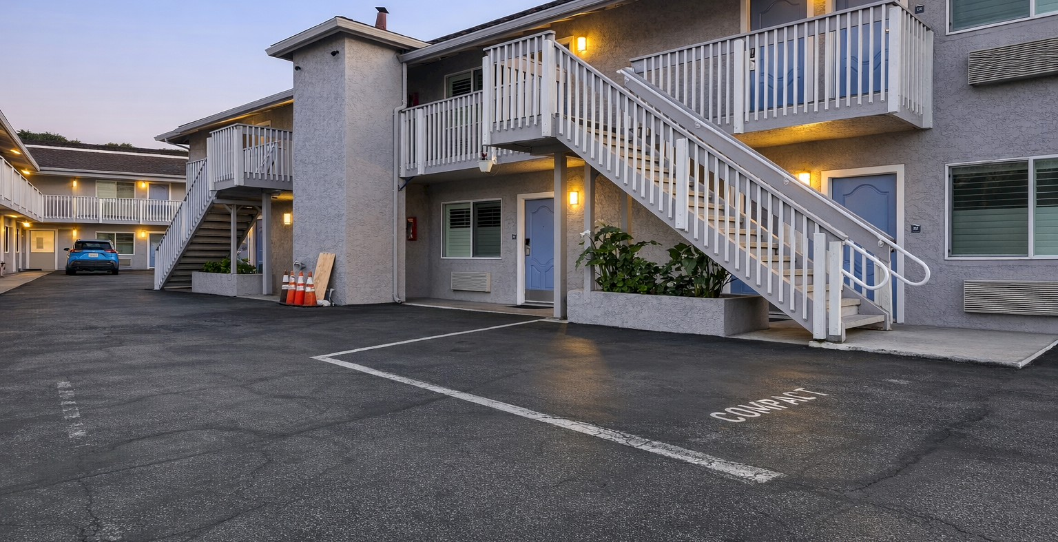 A two-story motel with exterior staircases, parking lot in front, warm lights at dusk, and a few balconies.
