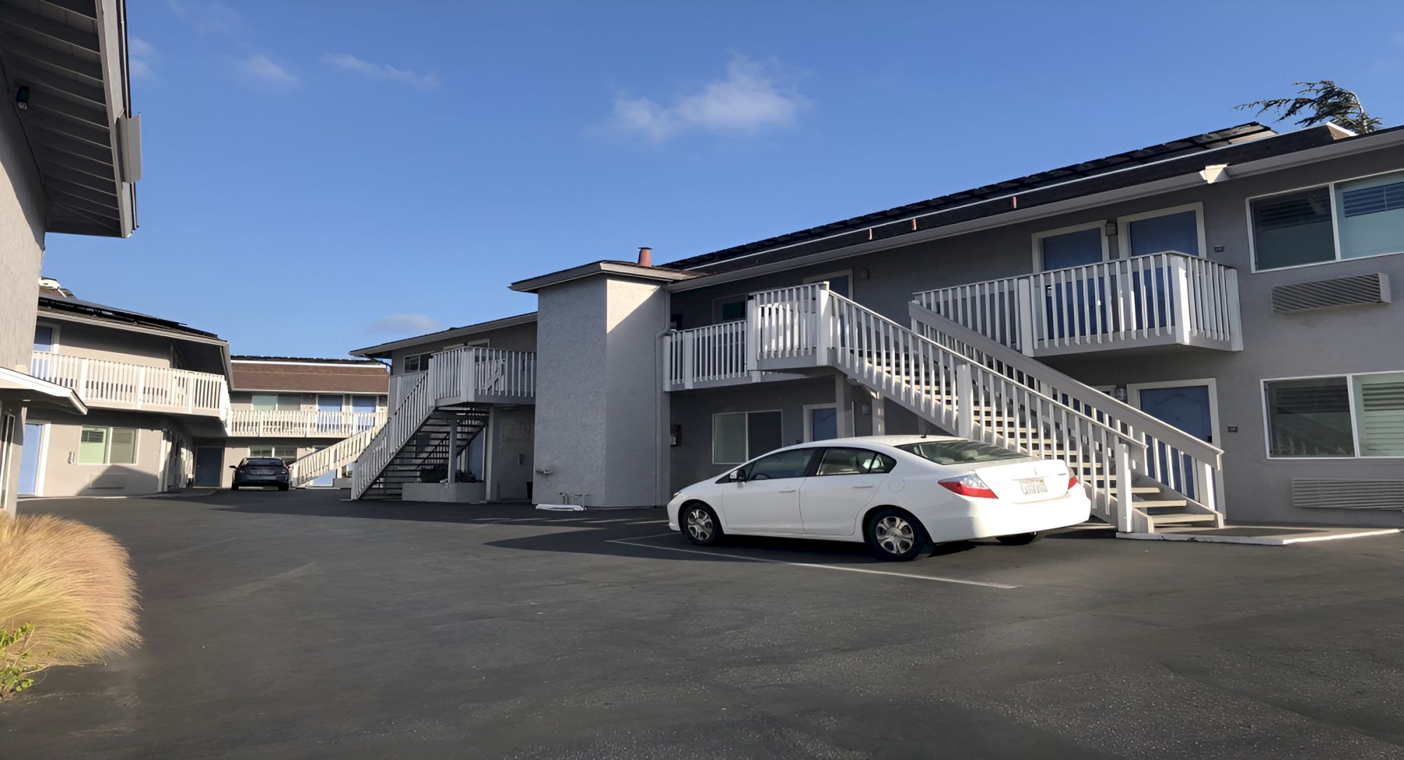 An exterior apartment complex with stairs to upper units, a white car parked in the lot, and a clear blue sky.