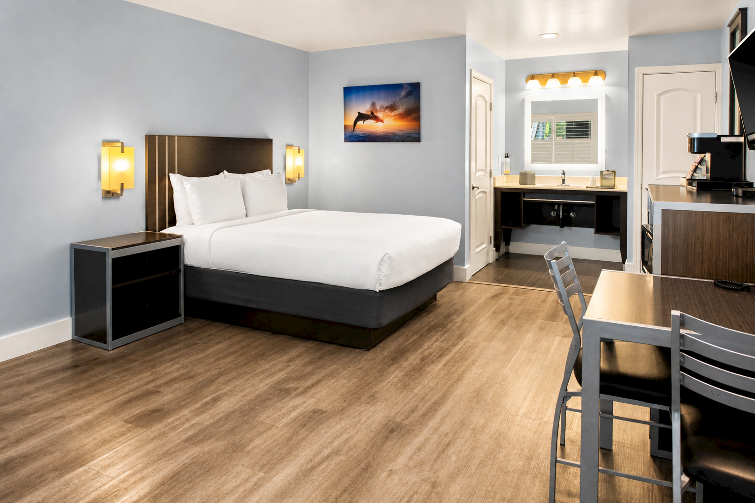 A modern hotel room with a king bed, nightstands, soft blue walls, wood flooring, a small desk area, and a kitchenette in the background, neat and bright.