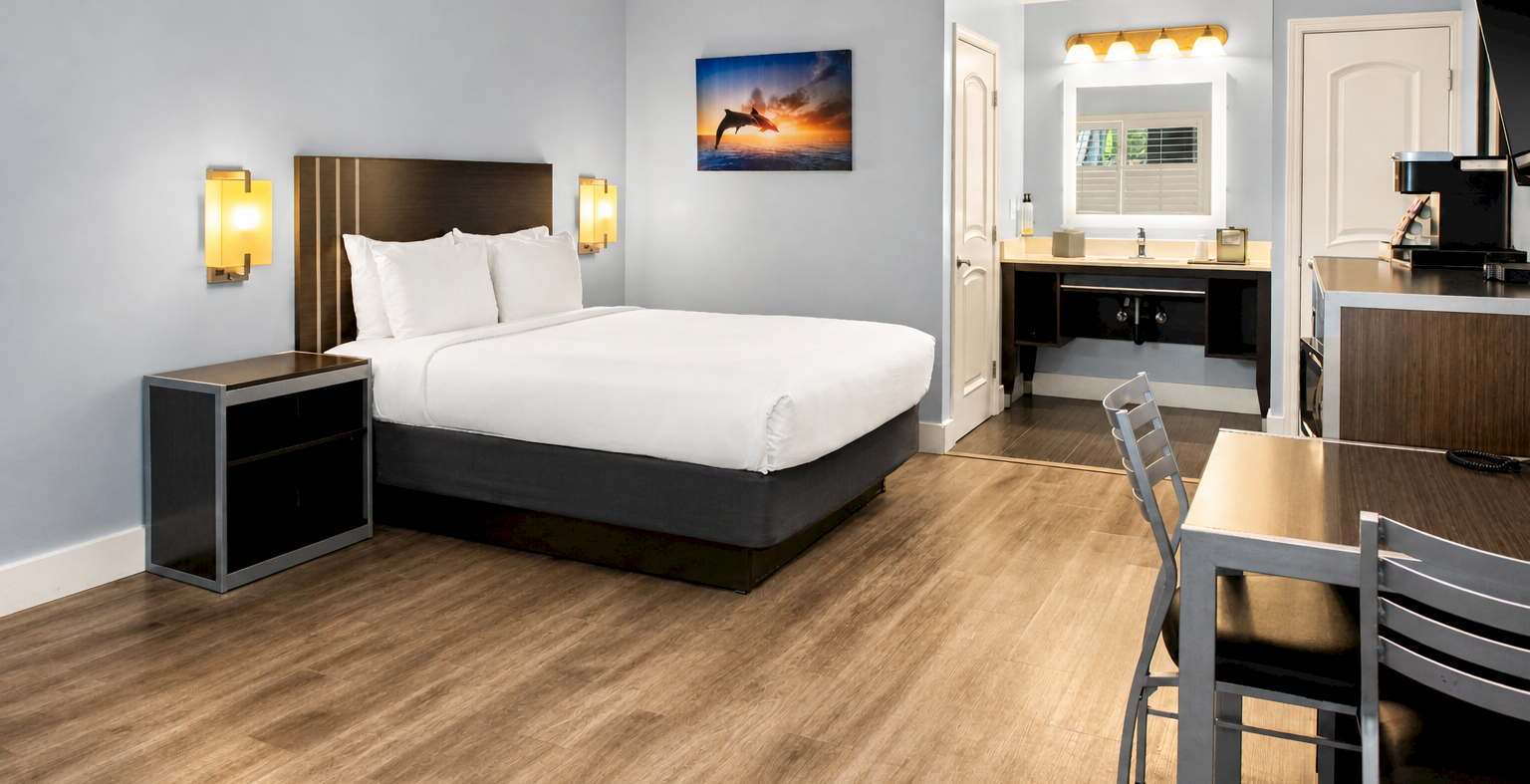 A modern hotel room with a king bed, nightstands, soft blue walls, wood flooring, a small desk area, and a kitchenette in the background, neat and bright.