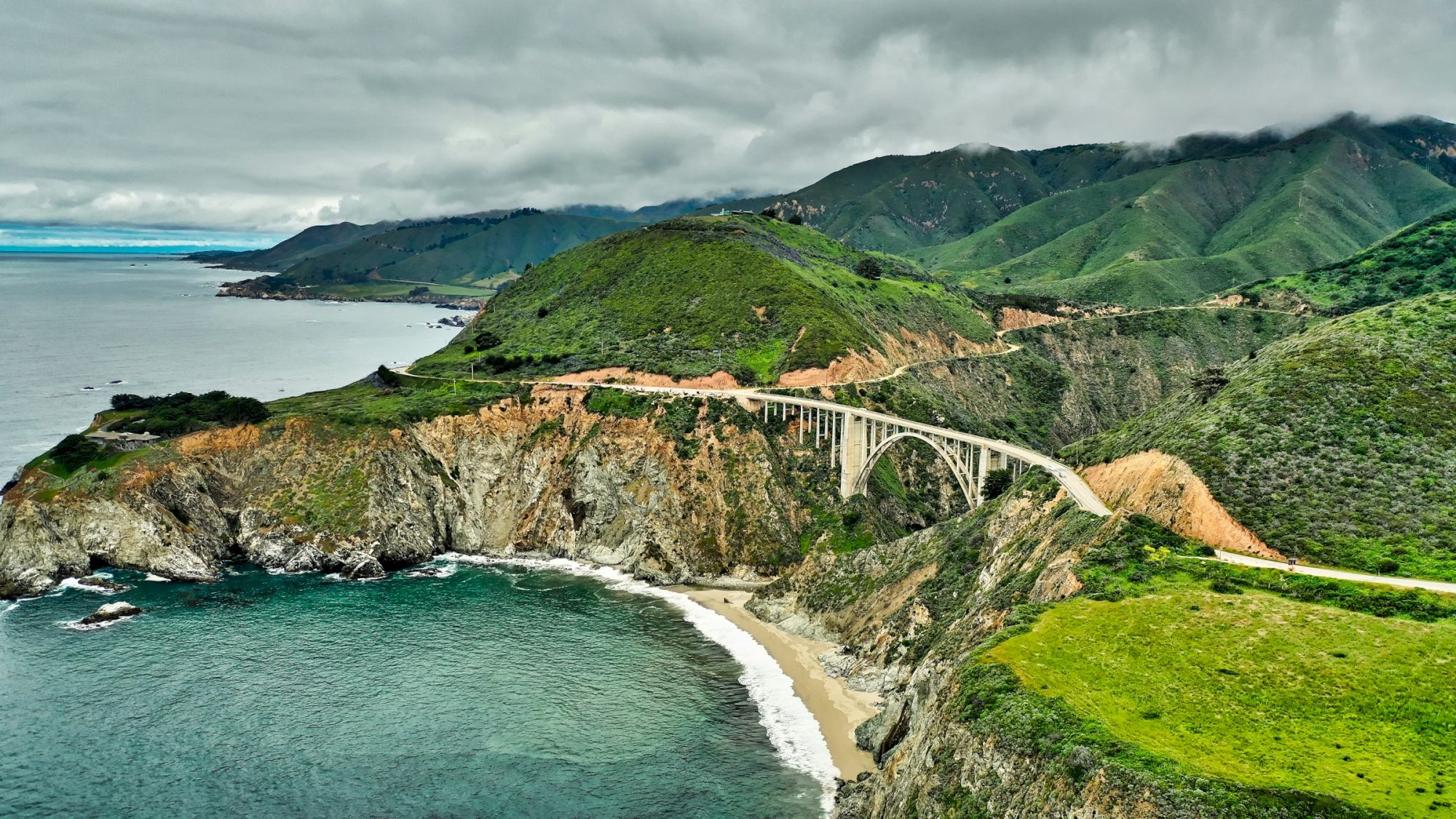 Coastal cliffs and a winding road along a bridge on a green hillside overlooking the ocean, with a rocky shoreline beneath.