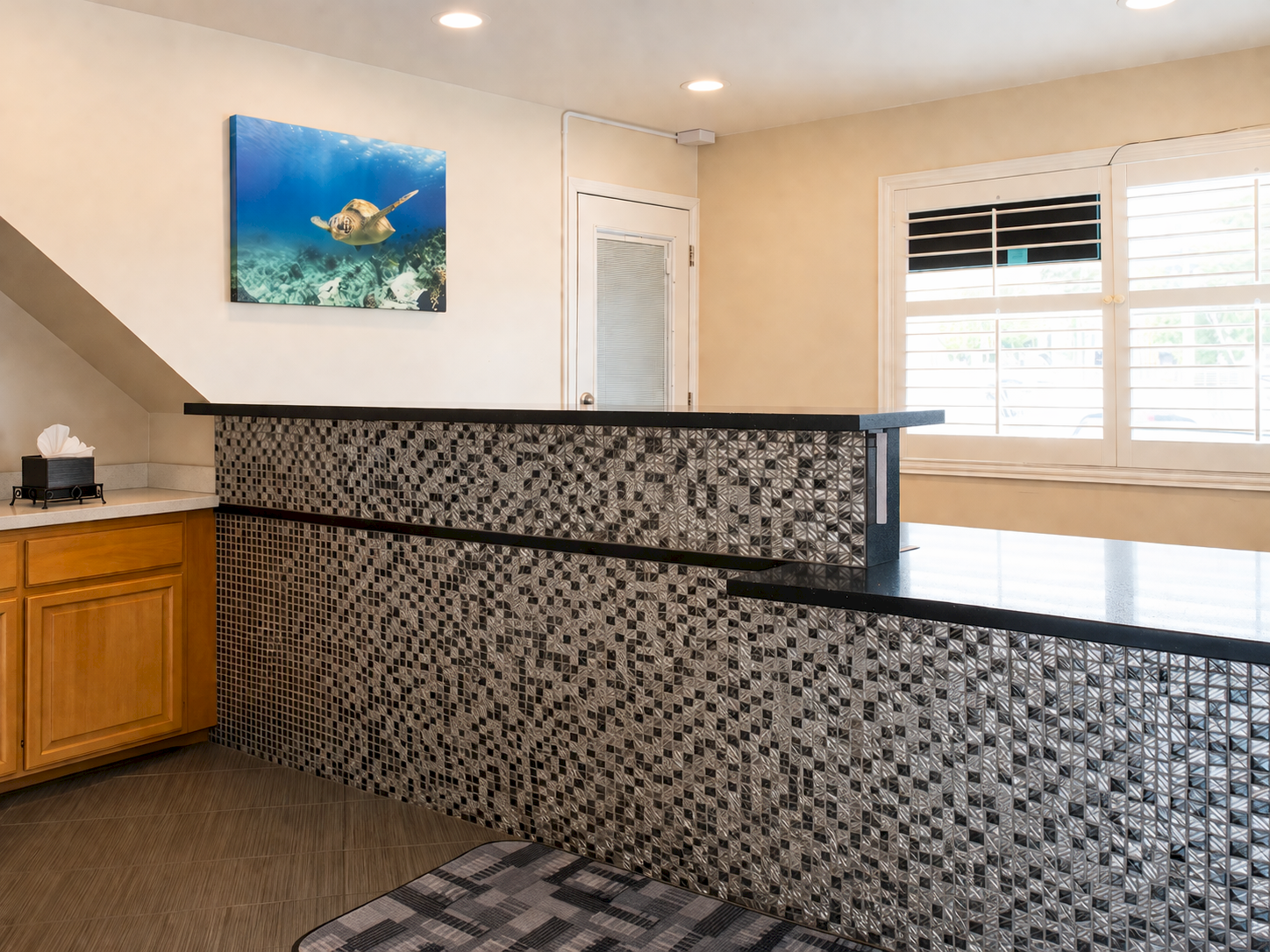 A bright bathroom with a tiled shower wall, wood vanity, seas turtle/ocean artwork, and a window with blinds and lights.