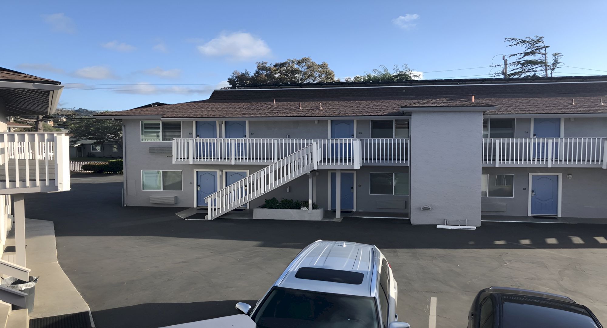 An apartment building with a central white staircase leading to upper units, a parking lot in front, and a few cars parked near the entrance.