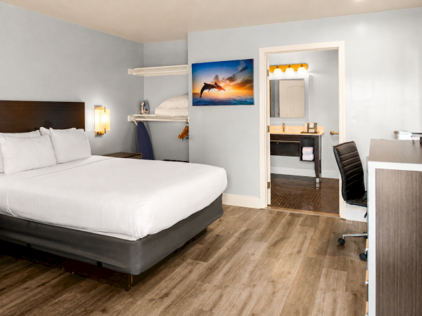 A modern hotel room with a king bed, wooden floor, desk, chair, wall art, open wardrobe, and a connected bathroom with a sink and mirror.