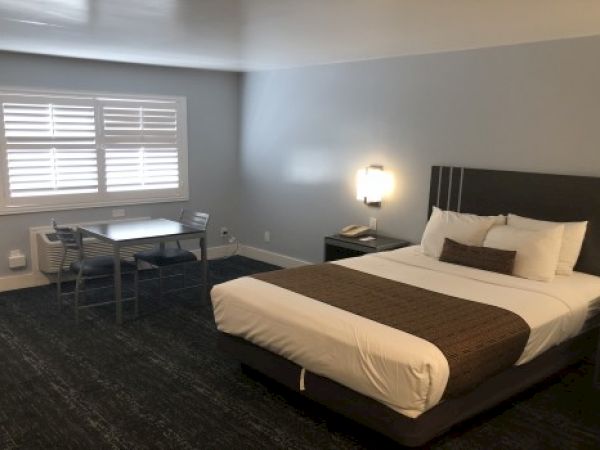A modern hotel room with a king bed, nightstands, a desk and chair, a window with blinds, and soft wall lighting, plus neutral decor.