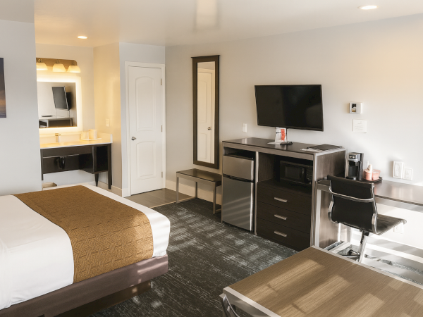 A modern hotel room with a bed, desk, TV, mini-fridge, mirror, and a small seating area; neutral tones and a clean, contemporary setup.
