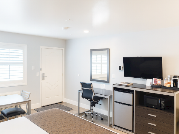 A hotel-style hotel room with a desk, chair, mini fridge, microwave, TV, drawers, a window, and a bed in the foreground.