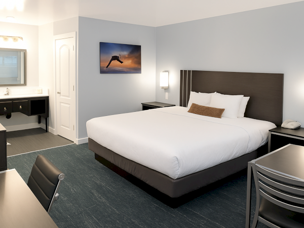 A clean hotel room with a king bed, two nightstands, a desk and chair, a wall-mounted lamp, a dresser, and a kitchenette area with a mirror.