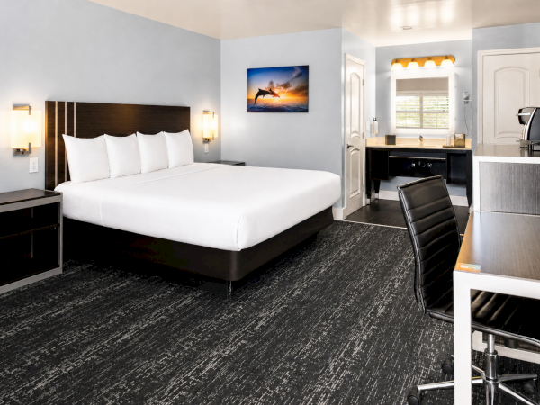 A modern hotel room with a king bed, two nightstands, a desk and chair, a flat-screen, mini-fridge/kitchenette area, and a small seating/coffee corner.