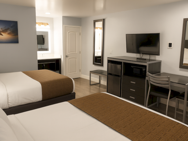 A hotel room with two beds, a desk and chairs, a wall-mounted TV, a dresser, mirror, and a small seating area near a door and kitchenette.