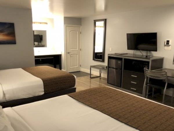 Two double beds in a hotel room with a desk, mini fridge, flat-screen TV, mirror, and a bathroom doorway nearby, neutral decor.