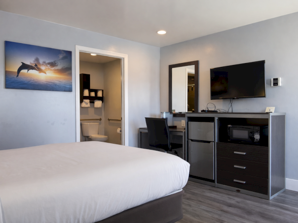 A hotel-style bedroom with a queen bed, blue-gray walls, a desk and chair, a wall-mounted TV, a mini-fridge and drawers, and a doorway to a bathroom&mdash;cozy and modern.