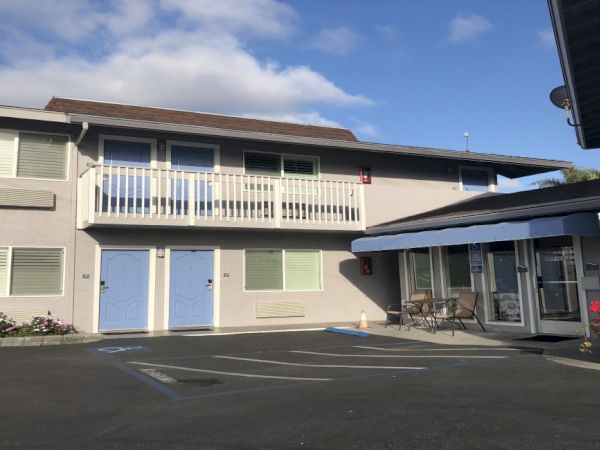 A two-story motel with light gray walls, blue doors, a white balcony, and a glass-wheeled patio area on the right, under a partly cloudy sky.