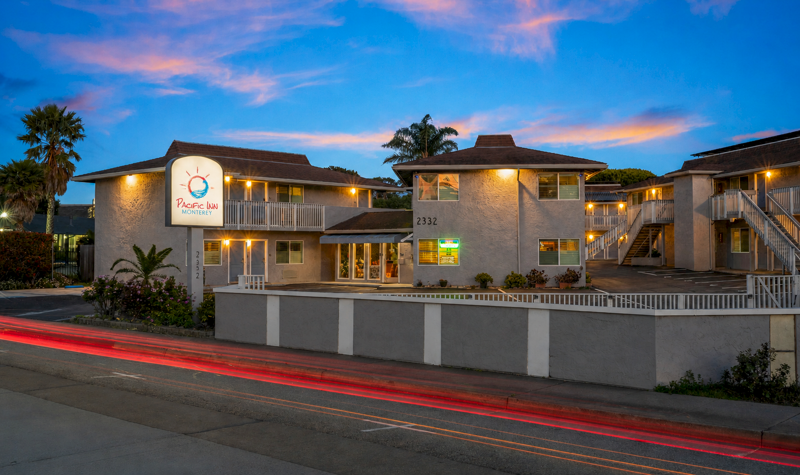 A Pacific Inn Monterey motel at dusk with palm trees, lit rooms, and red/white car light trails along the road.