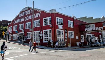 A red brick building labeled &ldquo;Monterey Canning Company&rdquo; on a sunny street, with people walking at a crosswalk. End.