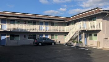 A two-story motel/apartment building with a central parking area and exterior stairs, under a partly cloudy sky.