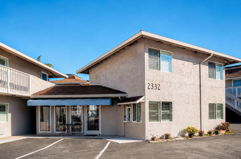 A single-story apartment building with a light beige exterior, numbered 2332, featuring large windows and a small entrance courtyard, under a clear blue sky.