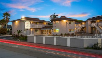 A small apartment complex with light-colored buildings, fenced yard, palm trees, and a street with red car light trails at dusk.