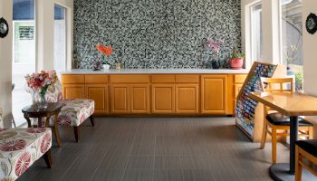 A bright reception area with a long wooden counter, mosaic tiled backsplash, and large windows. Seating includes floral-upholstered chairs, a small table with flowers, and a wooden high chair/stand near a counter. The floor is dark tile.