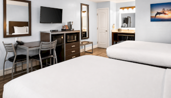 A clean hotel room with two neatly made beds, a desk and chair, a wall mirror, flat-screen TV, a mini-fridge, and a mirror reflecting a door and kitchenette.