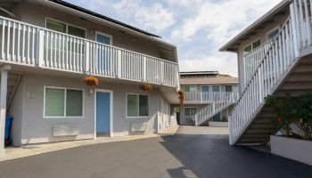 A two-story apartment complex with a central driveway, blue doors, white railings, hanging orange flowers, and external staircases on both sides.