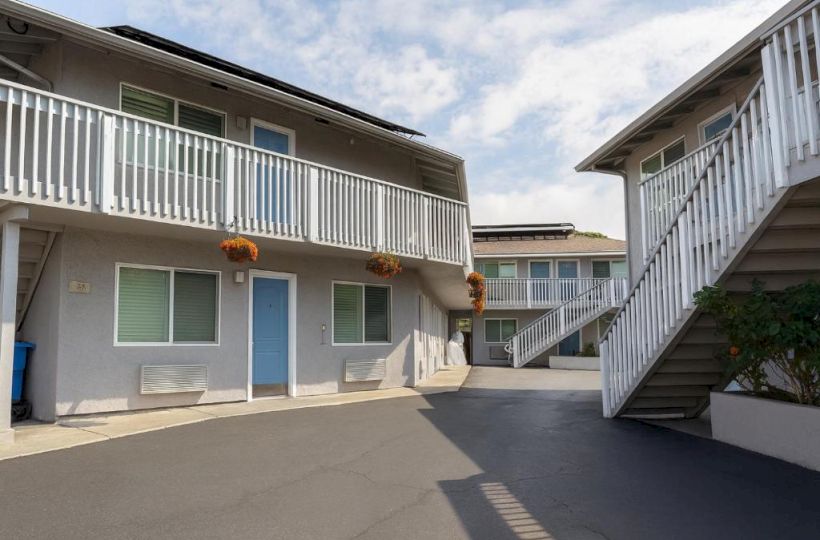 A two-story apartment complex with a central driveway, blue doors, white railings, hanging orange flowers, and external staircases on both sides.