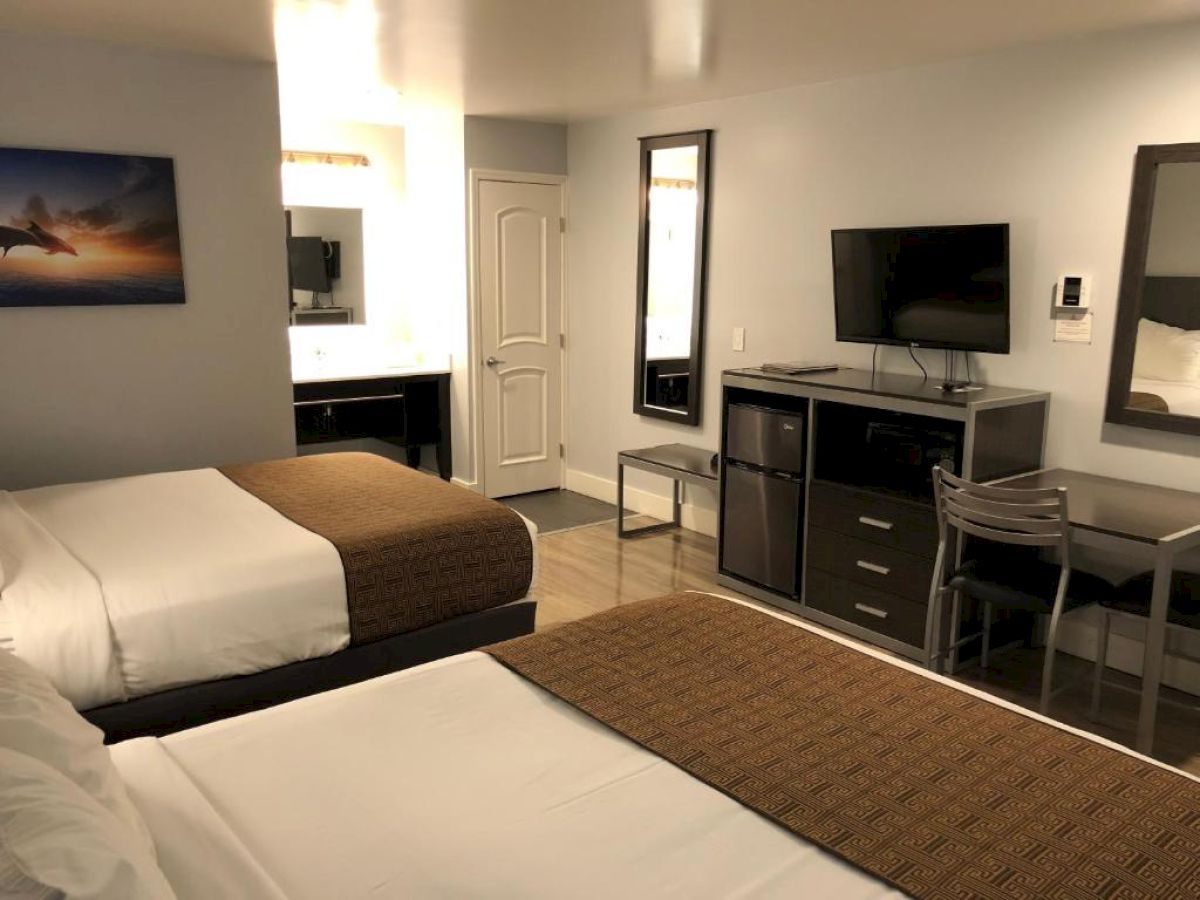 A hotel room with two beds, a dresser, mini-fridge, TV, desk, mirror, and a bathroom entry in the back, simple and tidy.