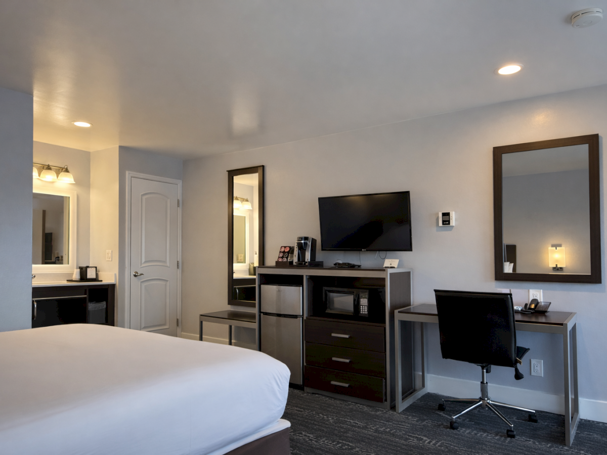 A hotel room with a large bed, desk with chair, wall mirror, TV on a dresser, mini-fridge and coffee station, and a doorway to a bathroom.