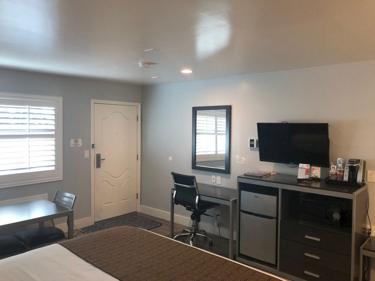 A hotel-style room with a bed, desk and chair, wall-mounted TV, mini fridge, coffee station, window blinds, and a small table near the door, end the sentence.
