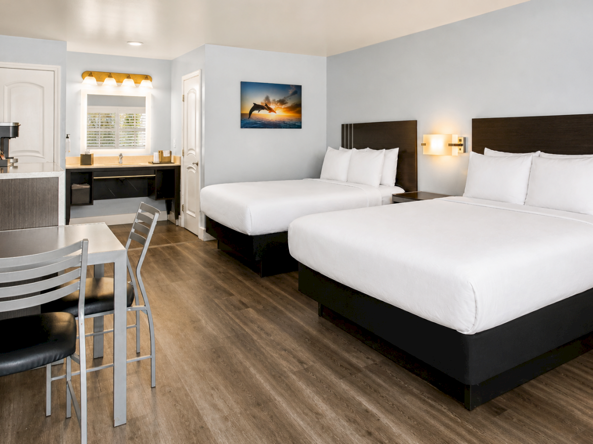 A tidy hotel room with two queen beds, a small desk and chair, a wall-mounted TV, a framed sunset picture, and a bright, airy feel.