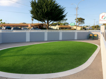A small circular grassy area enclosed by a light wall, with a curved concrete path, a tree, storefronts across the street, and a sign in the background.