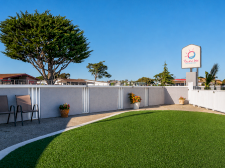 A sunny motel pool area with a curved grassy courtyard, white fence, lounge chairs, potted flowers, and a blue sky across a trim landscape.