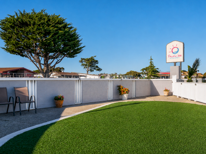 A sunny motel courtyard with a curved grassy lawn, white fence, lounge chairs, potted flowers, and a blue sky across a trim landscape.