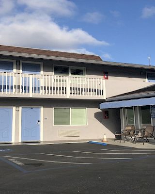 Residential motel-style building with a balcony, blue doors, shaded parking lot, and a glass-enclosed patio area on the right.