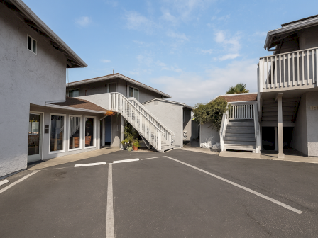A quiet motel-like courtyard with two-story white buildings, external stairs, apartments, and a paved center under a blue sky.