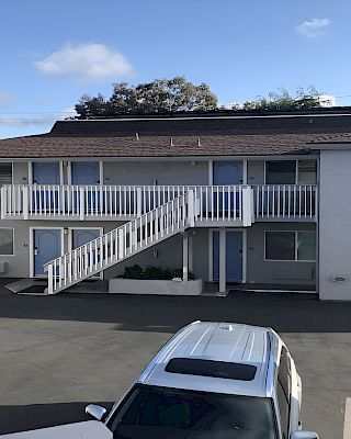 Two-story apartment building with exterior stairs, white railings, and a central walkway; a parking lot with a white SUV in front.