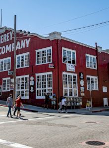 A colorful street scene with a red building on a corner, people walking, and parked cars by a blue sky, sunny day.