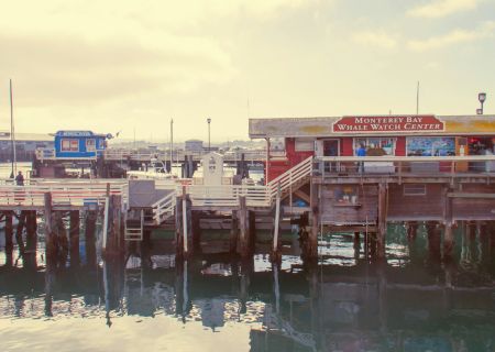 A wooden pier with a small building and signs, extending over calm water at a seaside town, under a soft sky.