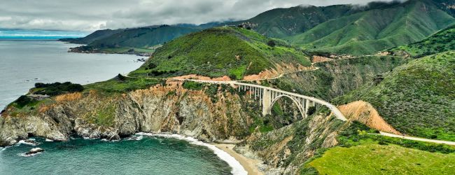 A coastal cliff road winds along turquoise sea, lush green hills, and dramatic shorelines under a bright sky.