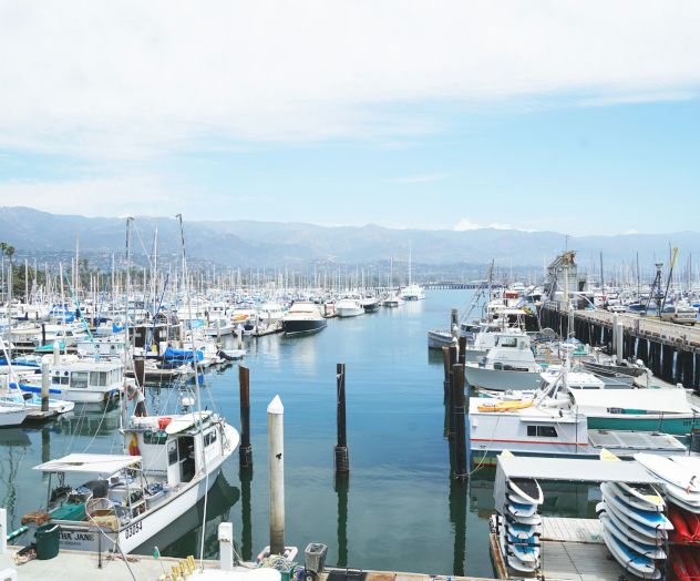 A busy marina with numerous boats and yachts docked along piers, calm water, and distant mountains under a partly cloudy sky.
