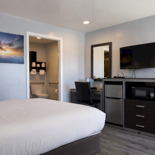 A modern hotel room with a white bed, wall art, a desk, a wall-mounted TV, and a small seating area.