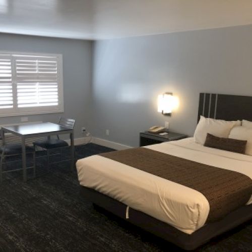 A modern hotel room with a king bed, dark headboard, white linens, brown throw, nightstands, a small desk and chair near a window with blinds.