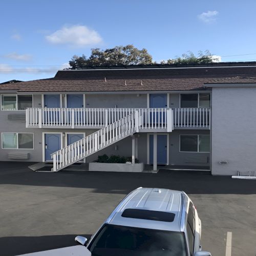 A two-story apartment building with white railings and exterior stairs, a parking lot in front, and a blue sky above.