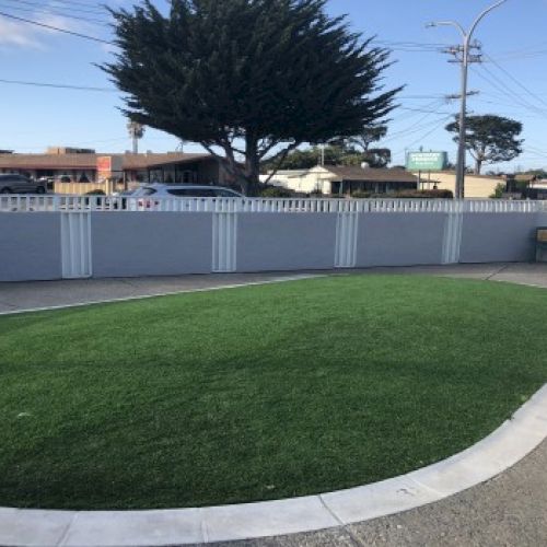 A circular grassy patch edged by a light concrete path, enclosed by a white fence, with a few trees and a street scene beyond.