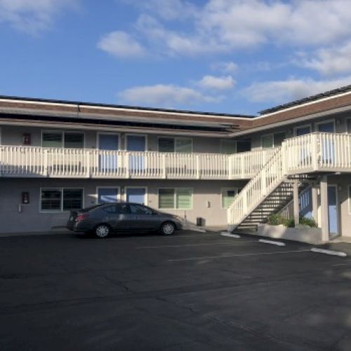 An outdoor apartment complex with a two-story building, white railings, a parking lot, a car, and a clear blue sky with a few clouds.