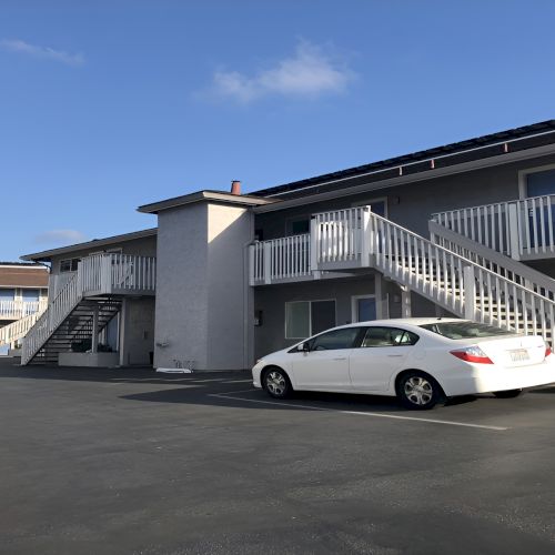 An apartment complex with exterior staircases, a white car parked in a lot, balconies, and a clear blue sky.