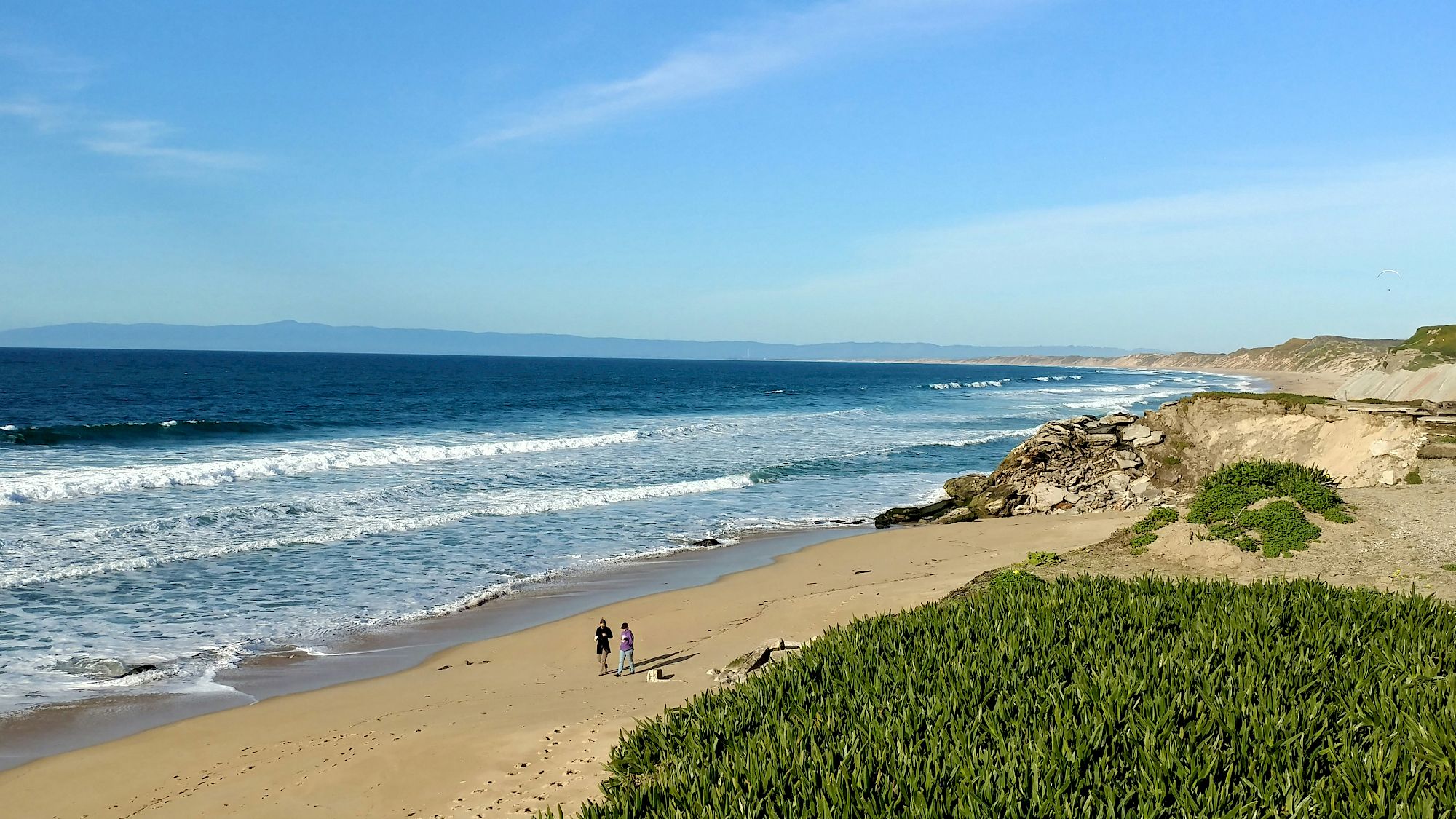A sandy beach with gentle waves, a rocky headland on the right, and a few people strolling near the shore under a clear blue sky.
