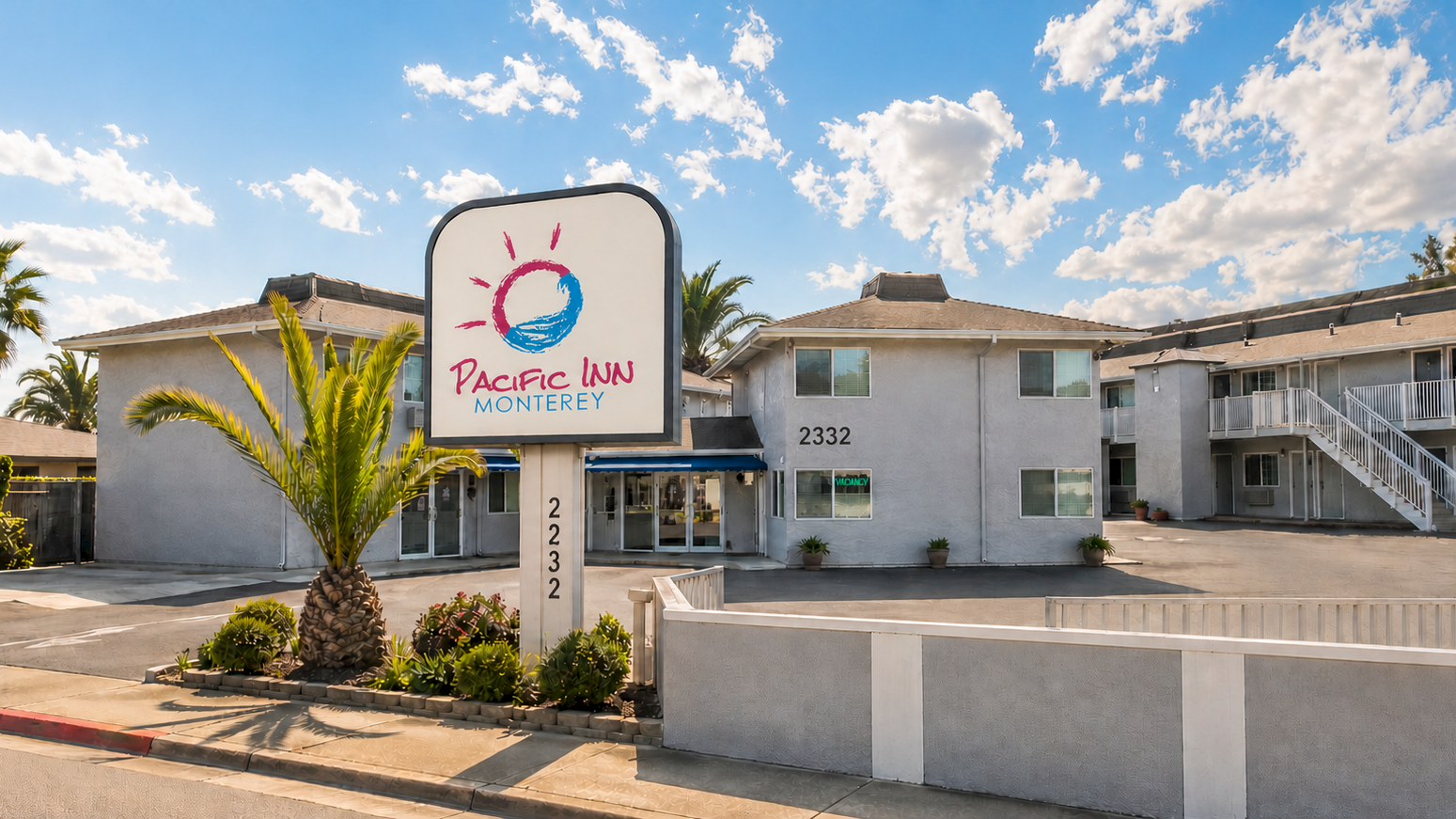 Pacific Inn Monterey: a sunny motel with a curved sign, palm trees, low-rise white buildings, and a small parking/driveway area.